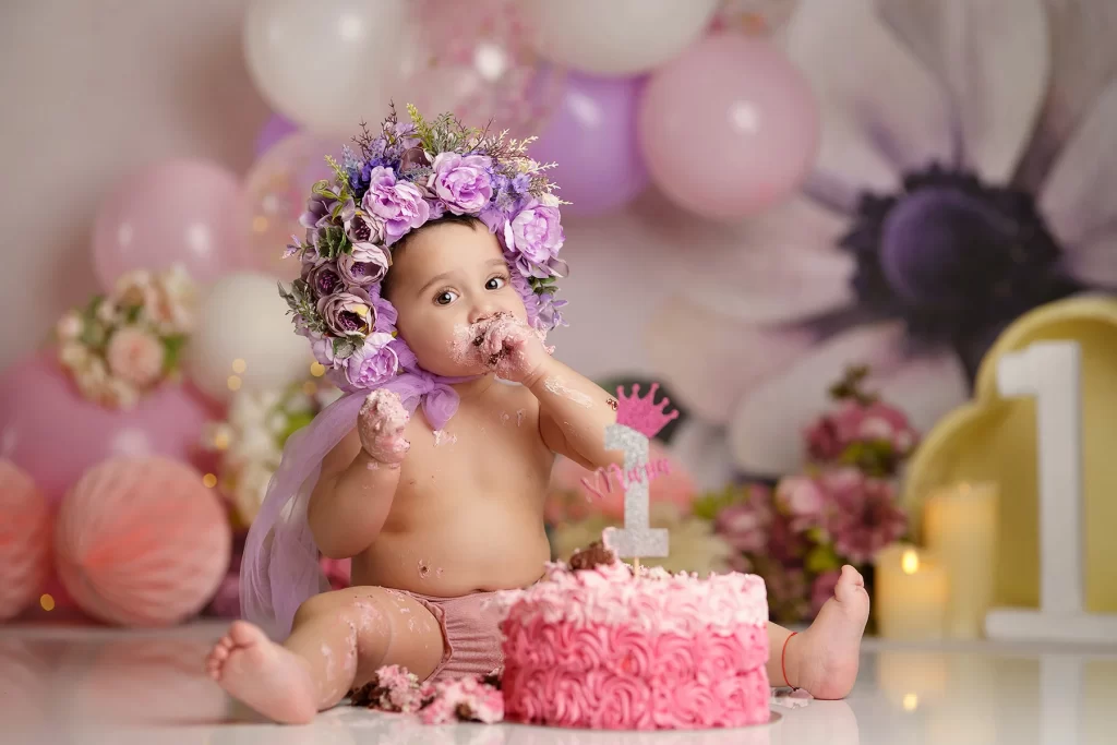 Cake smash photoshoot with a baby girl wearing a flower crown, enjoying her first birthday cake in a pink and pastel setup.