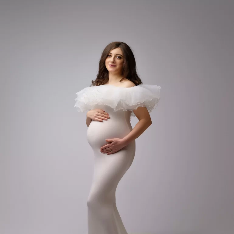 Maternity Photoshoot in North West London 50 Light‑gray backdrop; off‑shoulder white ruffled gown; gentle, proud pose.
