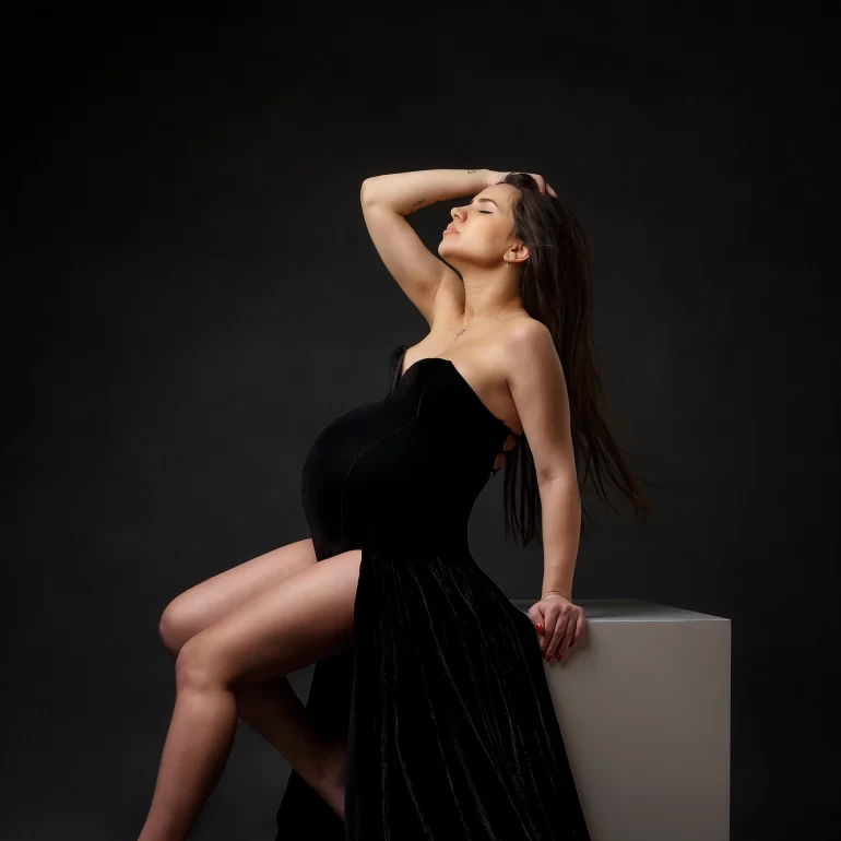 Maternity Photoshoot in North West London 51 Sleek black gown with high slit; seated on white block, confident pose.