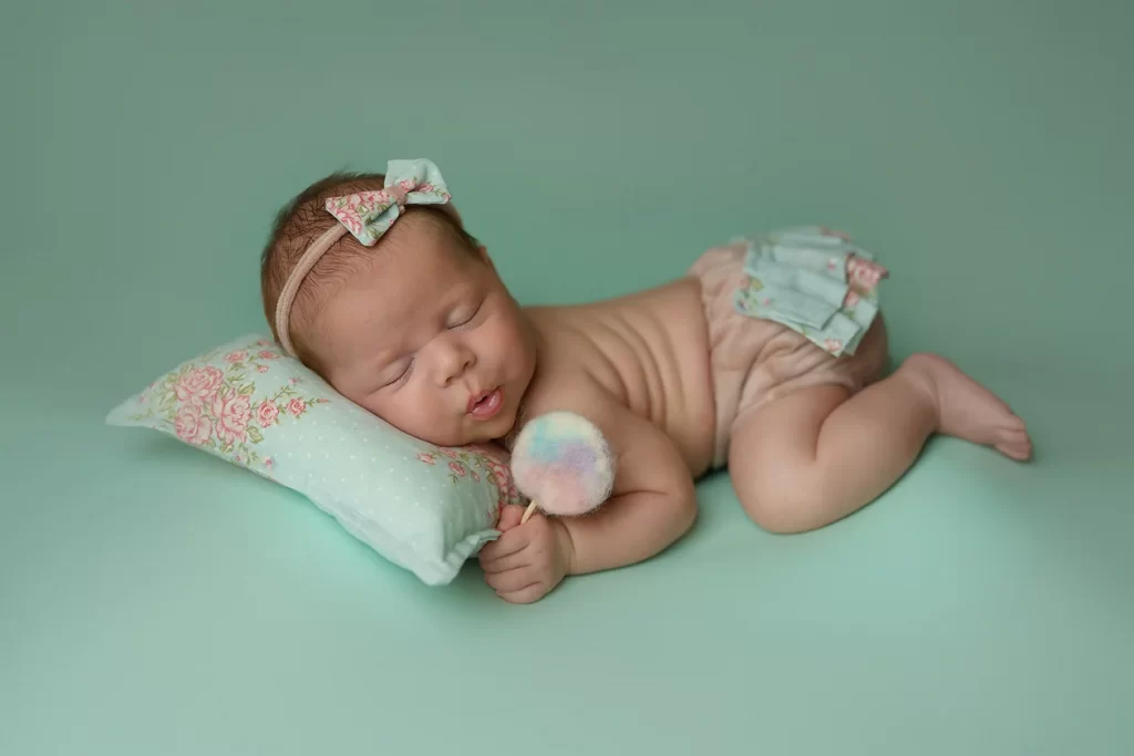 Newborn photoshoot with a baby girl sleeping on a mint background, holding a pastel wool pom-pom.