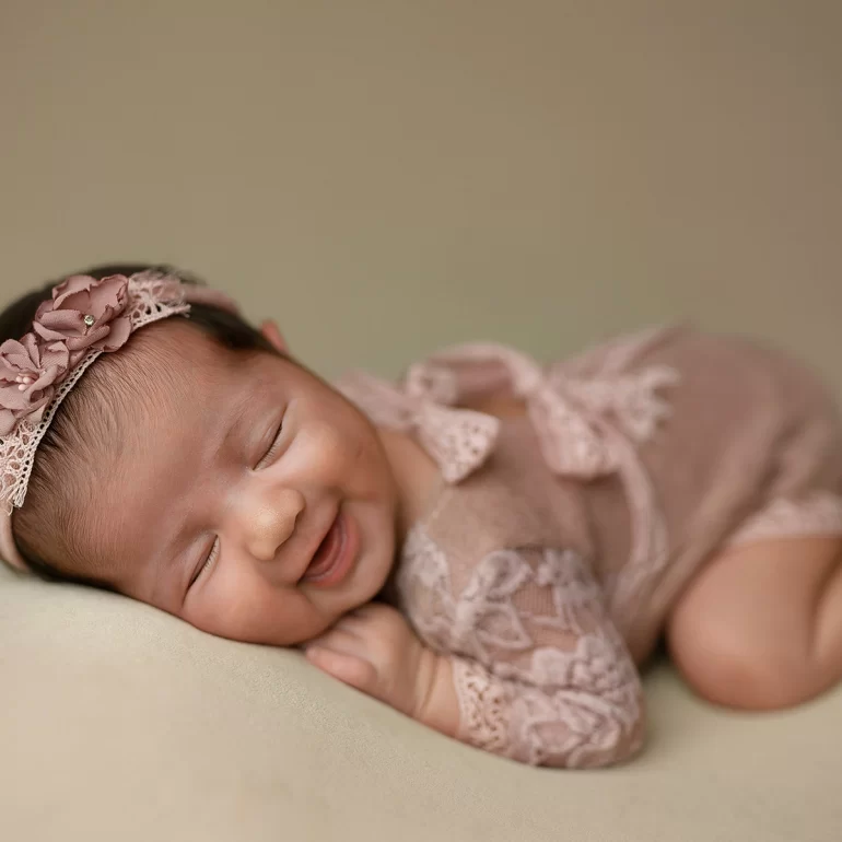Newborn Photoshoot in North West London 4 Smiling baby in blush lace with floral headband on soft beige backdrop.