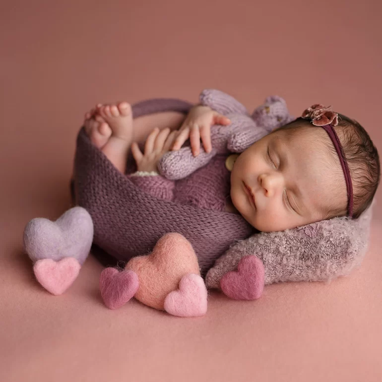 Newborn Photoshoot in North West London 18 Baby in purple wrap holding knit bear; pink backdrop with felt hearts.