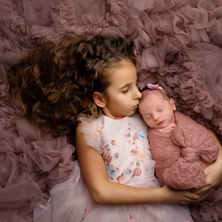 Newborn Photoshoot in North West London 19 Older sister holds swaddled baby in pink knit; ruffled mauve backdrop.