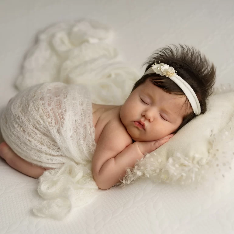 Newborn Photoshoot in North West London 5 Baby on cream pillow with sheer lace wrap and white floral headband; airy whites.