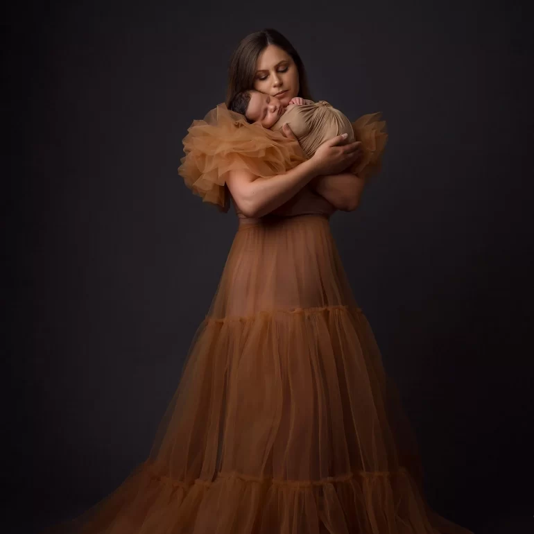 Newborn Photoshoot in North West London 23 Mother in sheer brown dress cradling swaddled baby; soft, warm tones.