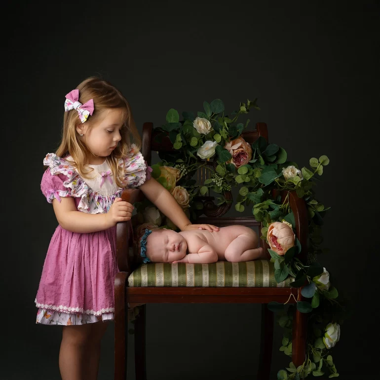 Newborn Photoshoot in North West London 24 Young sister touches sleeping baby on green striped chair with florals.