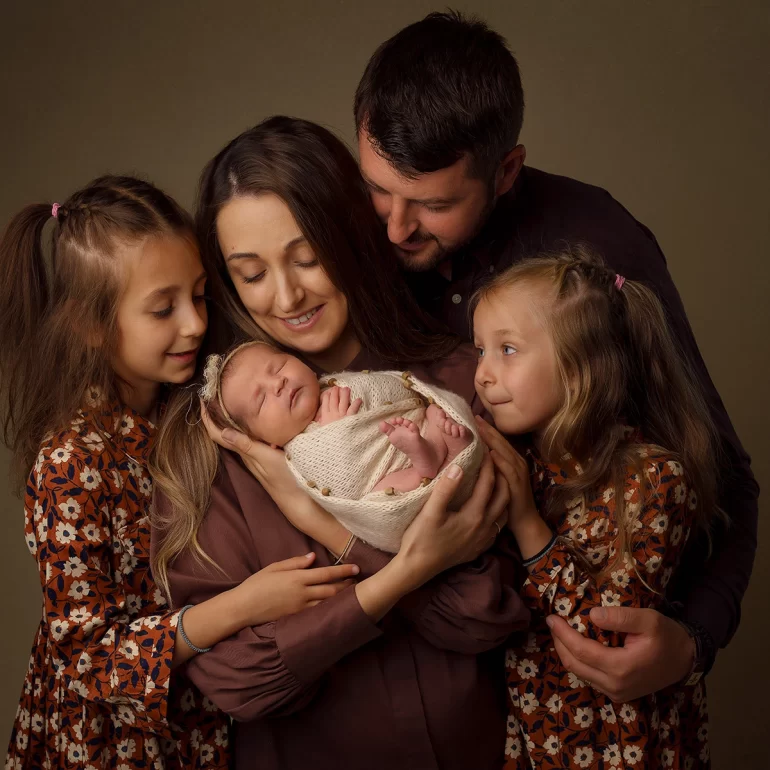 Newborn Photoshoot in North West London 25 Parents hold swaddled baby; two daughters in floral dresses embrace; warm light.