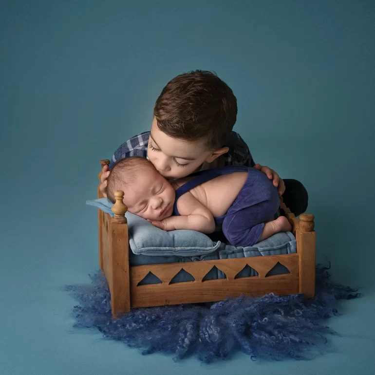 Newborn Photoshoot in North West London 27 Older sibling kisses baby on mini bed; soft blue tones and textures.