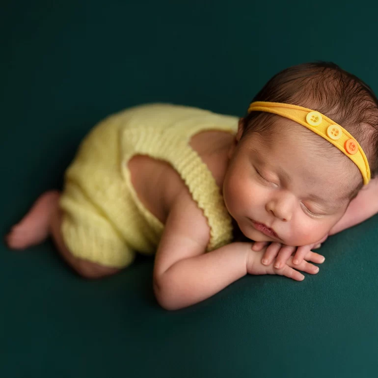 Newborn Photoshoot in North West London 28 Baby in yellow knit romper and headband on rich green backdrop.