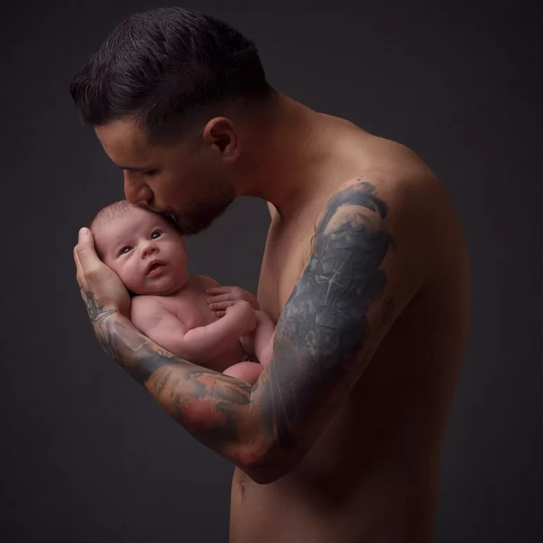 Newborn Photoshoot in North West London 29 Tattooed father kisses baby’s head against dark studio; tender moment.