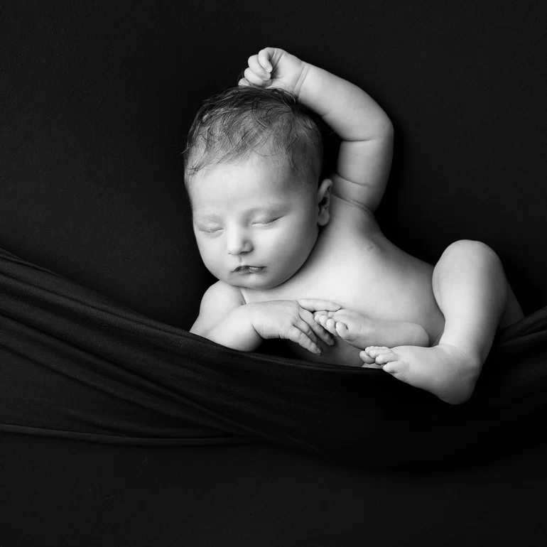 Newborn Photoshoot in North West London 32 B&W baby in suspended wrap, one arm raised; calm, dark background.