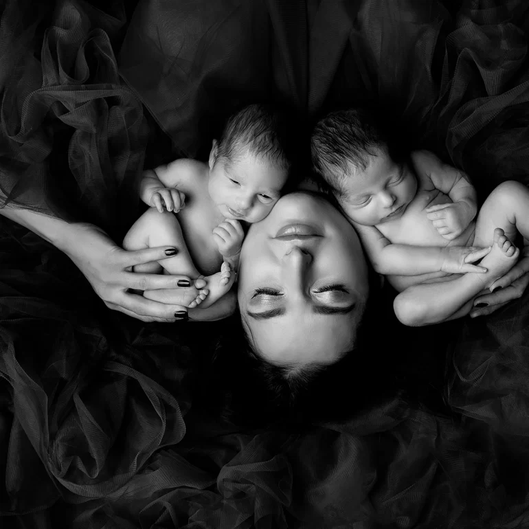Newborn Photoshoot in North West London 35 B&W mother lying back holding twin babies; soft tulle surrounds.