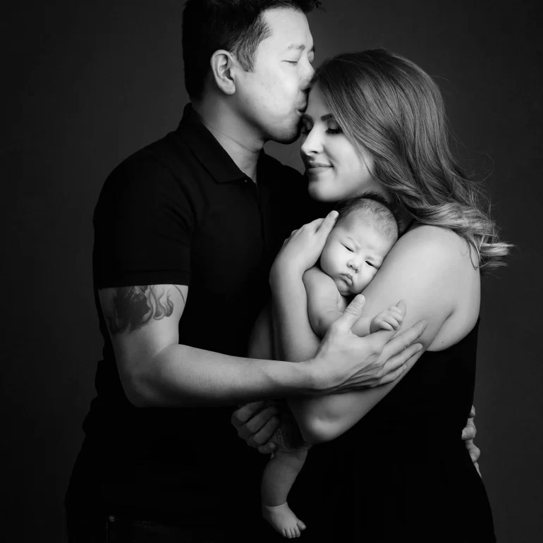 Newborn Photoshoot in North West London 36 B&W family: father forehead kiss as mother holds baby; warm, tender.