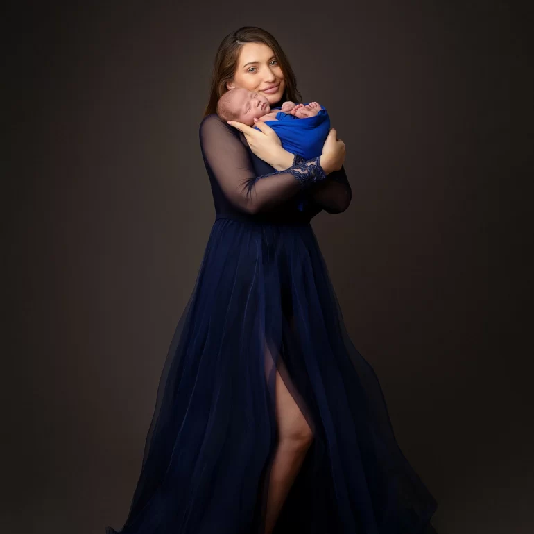 Newborn Photoshoot in North West London 37 Mother in navy gown cradling swaddled baby; dark studio backdrop.