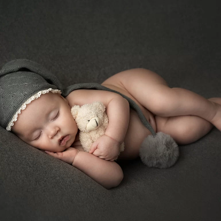 Newborn Photoshoot in North West London 38 Baby on dark surface with grey lace‑trim hat, holding small teddy.