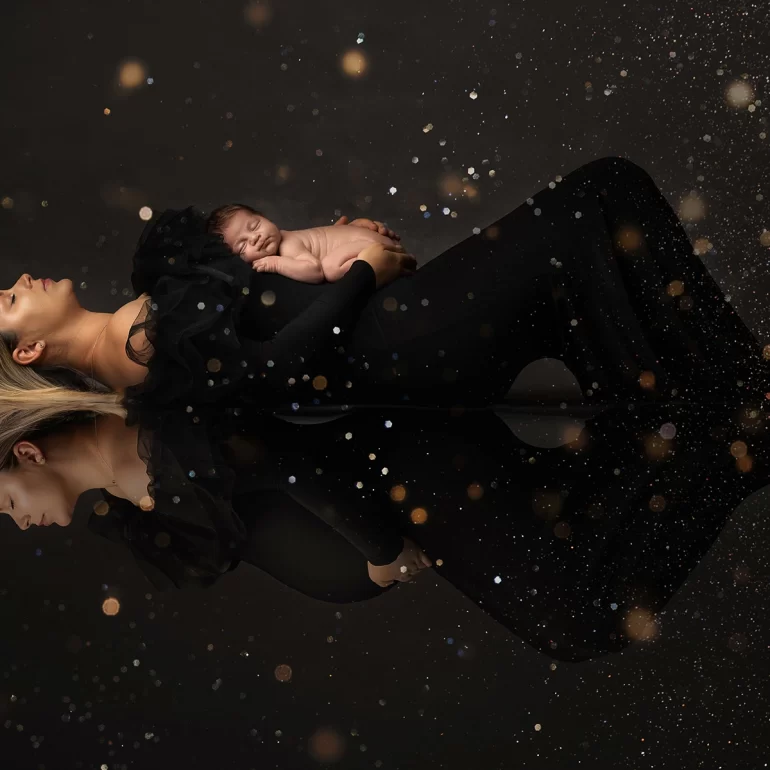 Newborn Photoshoot in North West London 39 Mother in black gown holding baby; golden bokeh and mirrored reflection.