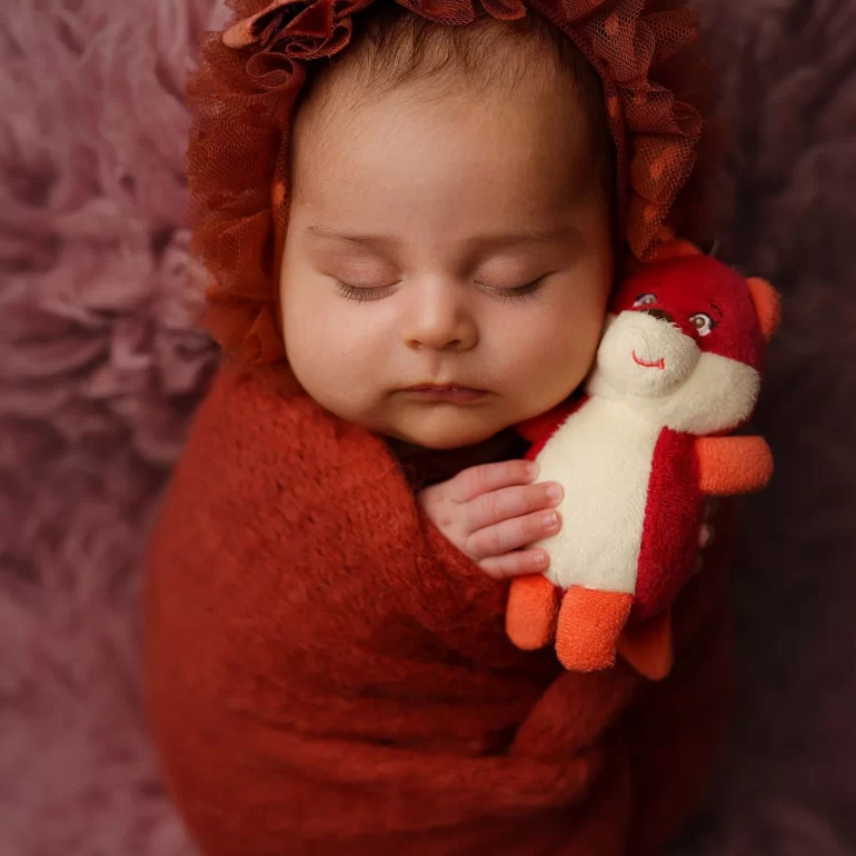 Newborn Photoshoot in North West London 40 Baby in red swaddle with lace bear‑ear bonnet; holding tiny toy; purple backdrop.