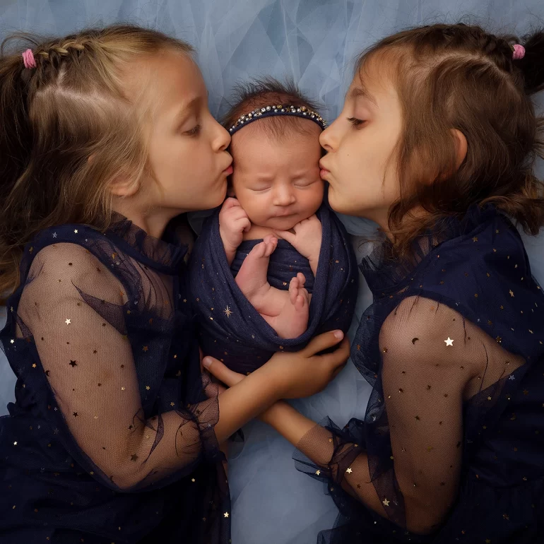 Newborn Photoshoot in North West London 44 Two sisters in navy with golden stars kiss swaddled sibling on tulle.