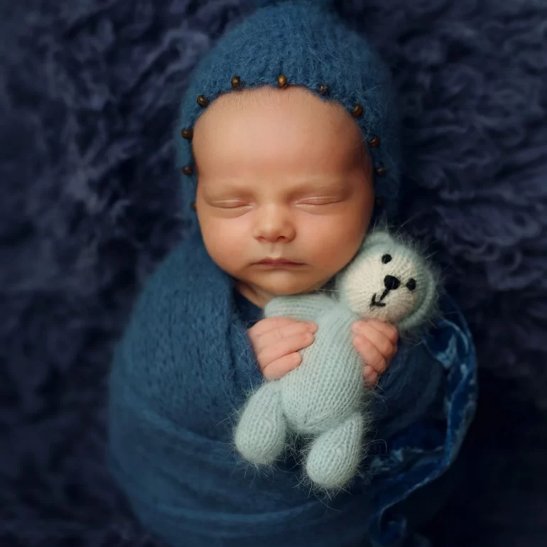 Newborn Photoshoot in North West London 45 Baby in blue swaddle and beaded bonnet holding tiny knit teddy; fluffy backdrop.