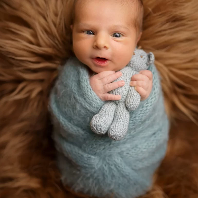 Newborn Photoshoot in North West London 46 Wide‑eyed baby in blue knit with teddy on brown fur; warm, inviting.