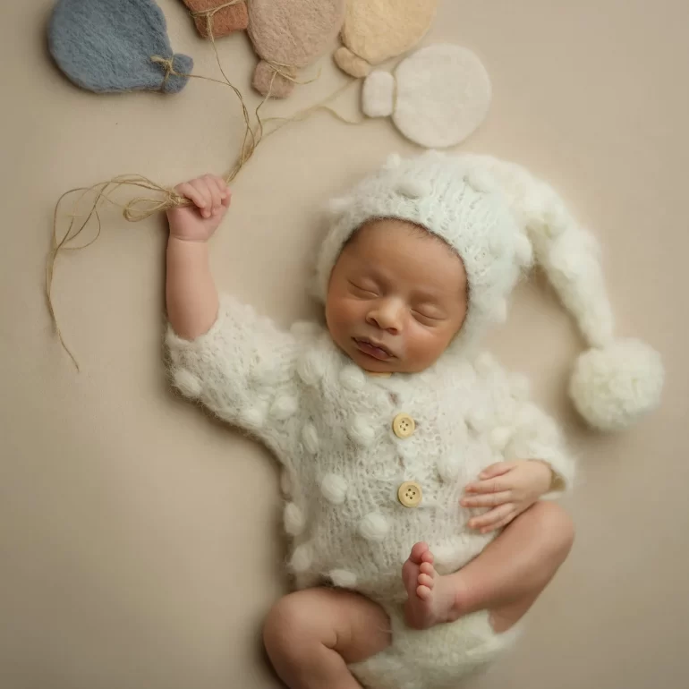 Newborn Photoshoot in North West London 51 Baby in white knit with pom‑poms holding felt balloon strings; soft beige set.