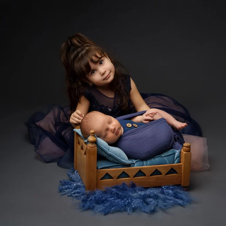 Newborn Photoshoot in North West London 52 Girl in navy poses by baby on carved mini bed; rich, dark backdrop.