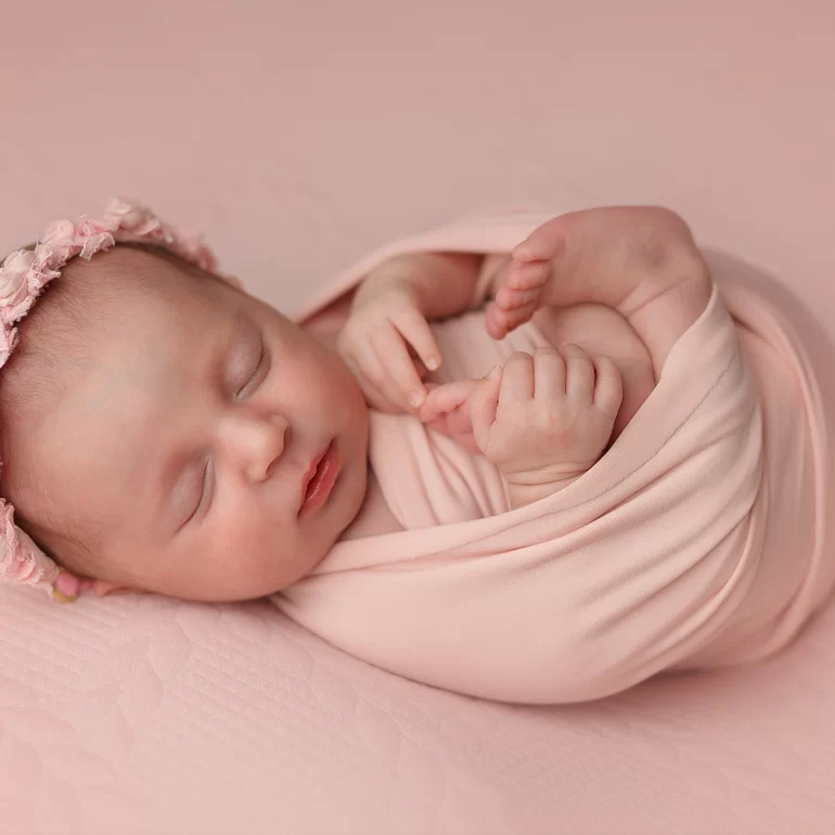 Newborn Photoshoot in North West London 58 Baby in pastel pink swaddle with floral headband on quilted backdrop.