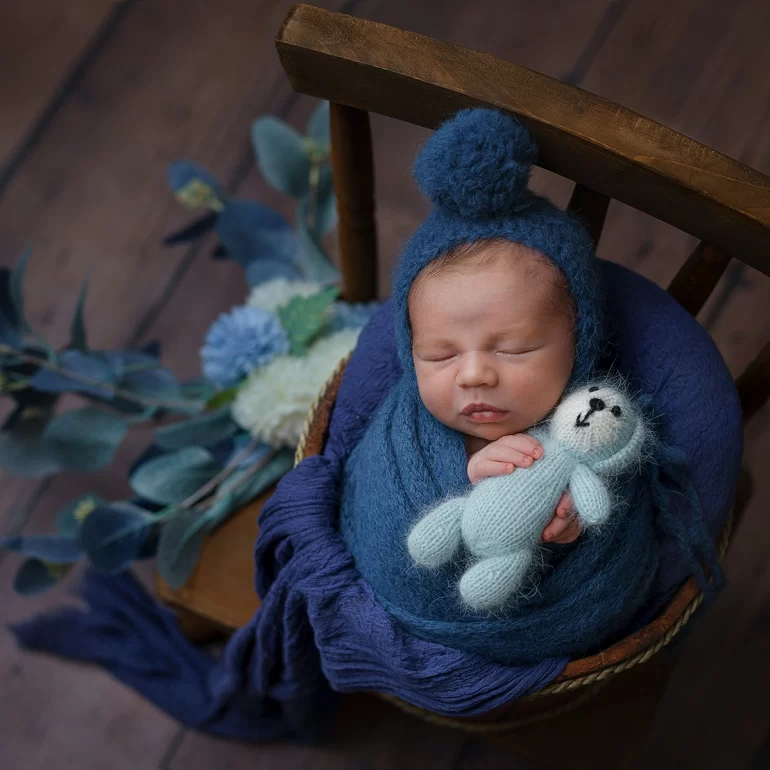 Newborn Photoshoot in North West London 59 Baby in navy wrap and pom‑pom bonnet seated in rustic chair with florals.