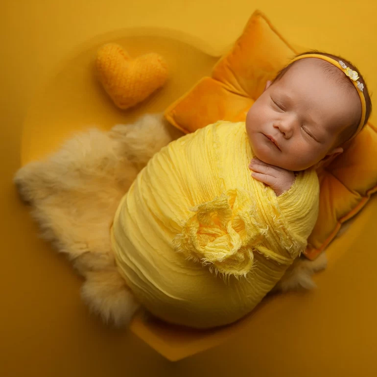 Newborn Photoshoot in North West London 61 Baby in bright yellow wrap with heart prop on warm golden set; cheerful mood.