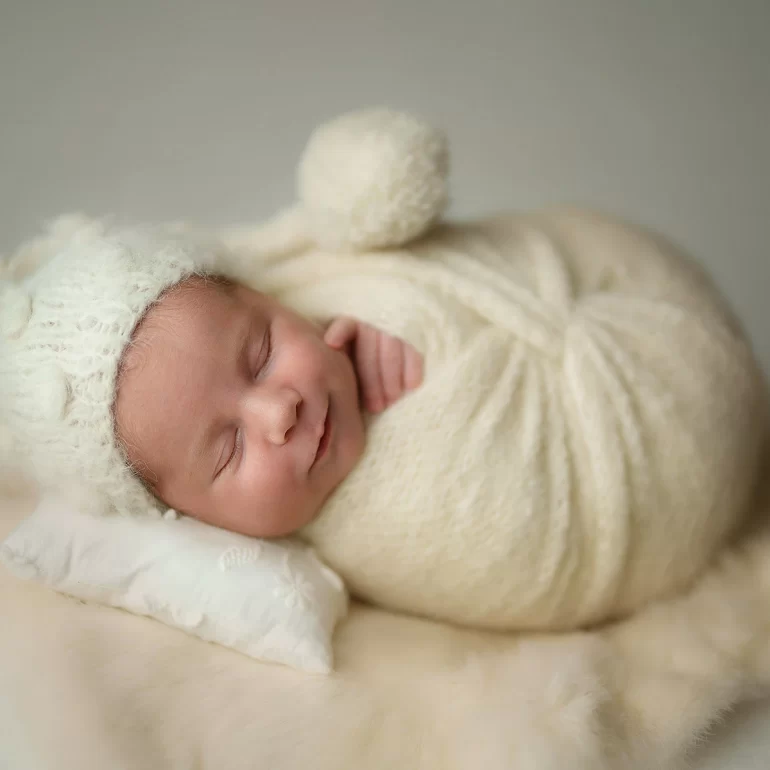 Newborn Photoshoot in North West London 63 Baby in ivory knit with pom‑pom hat on cream fur and embroidered pillow.