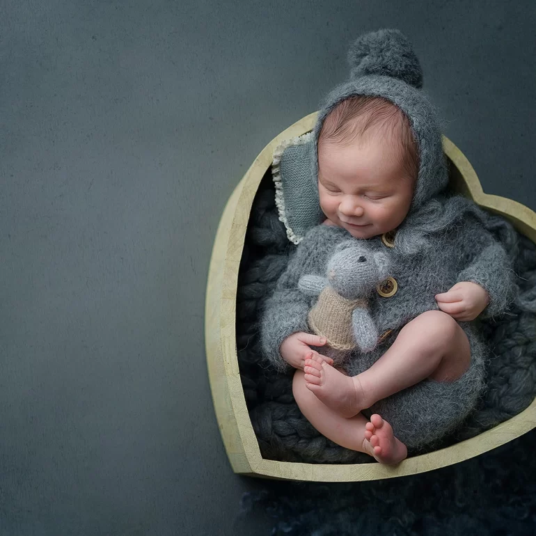 Newborn Photoshoot in North West London 66 Smiling baby in grey knit with bonnet and toy in heart‑shaped wooden bowl.