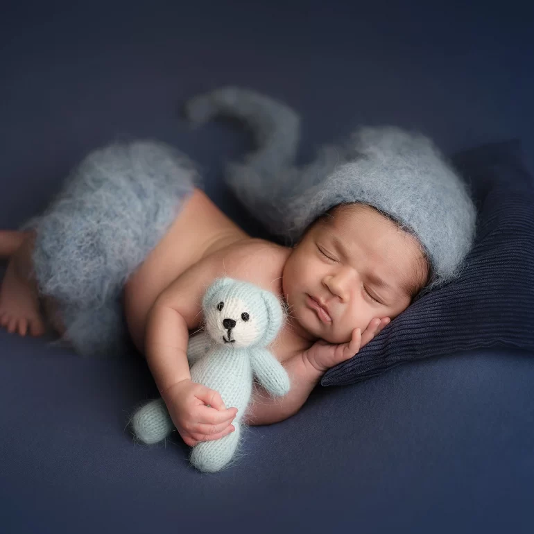 Newborn Photoshoot in North West London 68 Baby in blue knit and whimsical hat cuddling teddy on plush navy pillow.