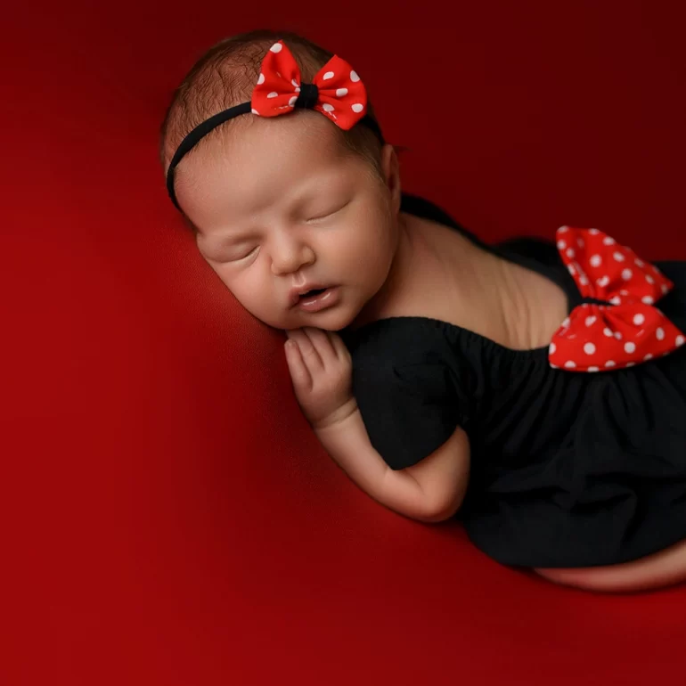 Newborn Photoshoot in North West London 71 Baby on red backdrop in black outfit with red polka‑dot bows and headband.