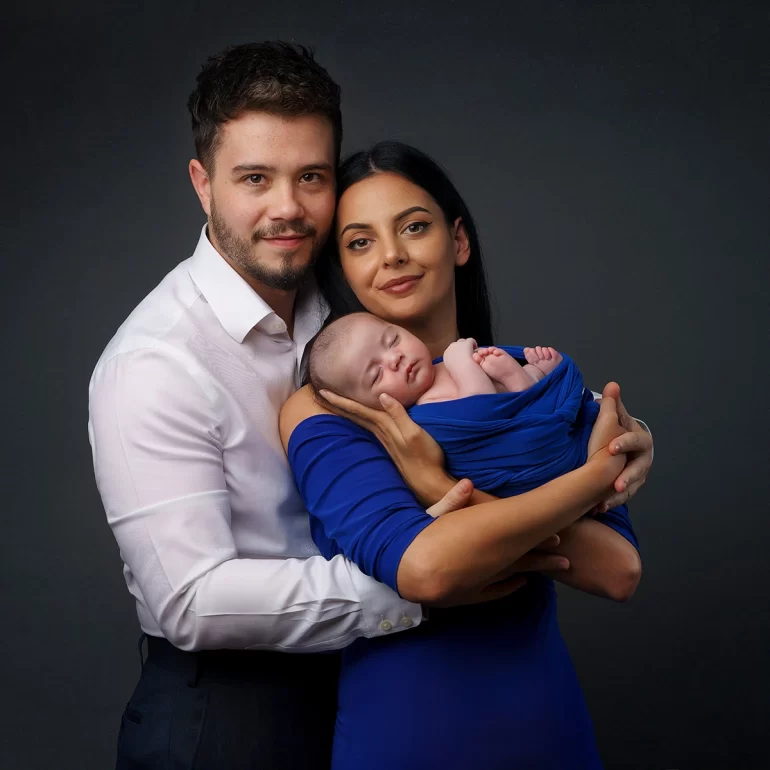 Newborn Photoshoot in North West London 78 Father in white, mother in royal blue holding baby in matching swaddle; elegant dark set.