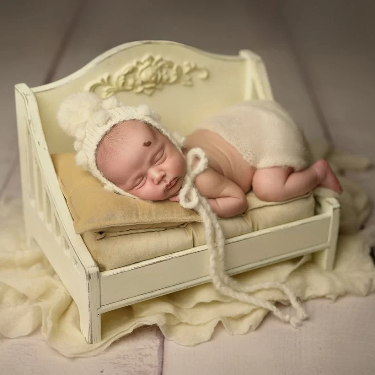 Newborn Photoshoot in North West London 79 Baby on cream mini bed in ivory bonnet with pom‑poms; warm neutrals.
