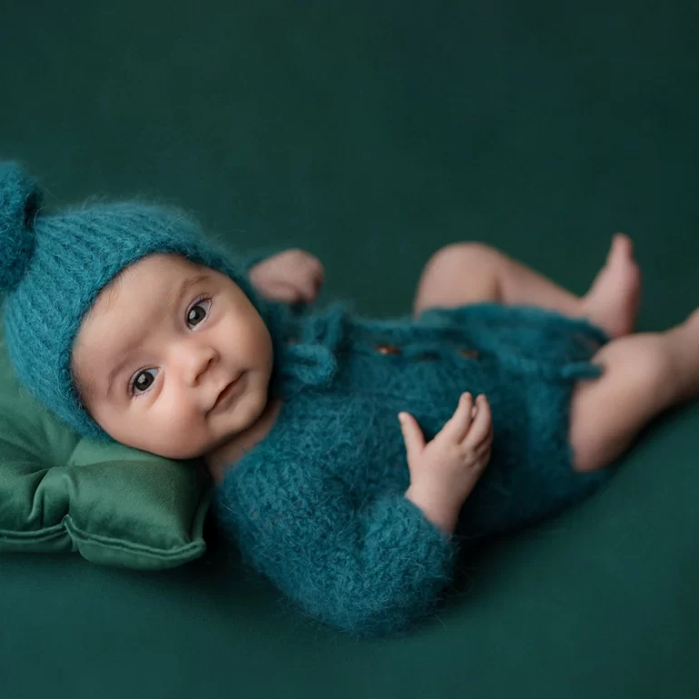 Newborn Photoshoot in North West London 80 Baby in teal knit romper and pom‑pom hat on plush green cushion; dark green set.