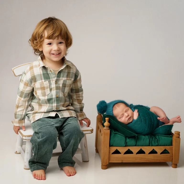Newborn Photoshoot in North West London 81 Smiling toddler on white chair beside baby on mini bed in teal knit; warm bond.