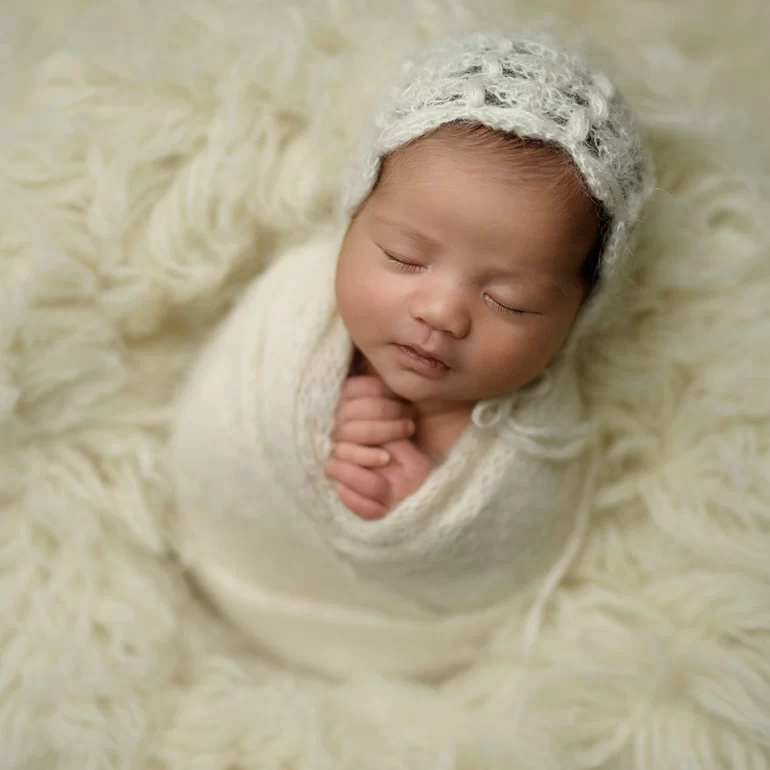 Newborn Photoshoot in North West London 84 Baby in cream wrap with knit bonnet on fluffy cream backdrop; hands clasped.