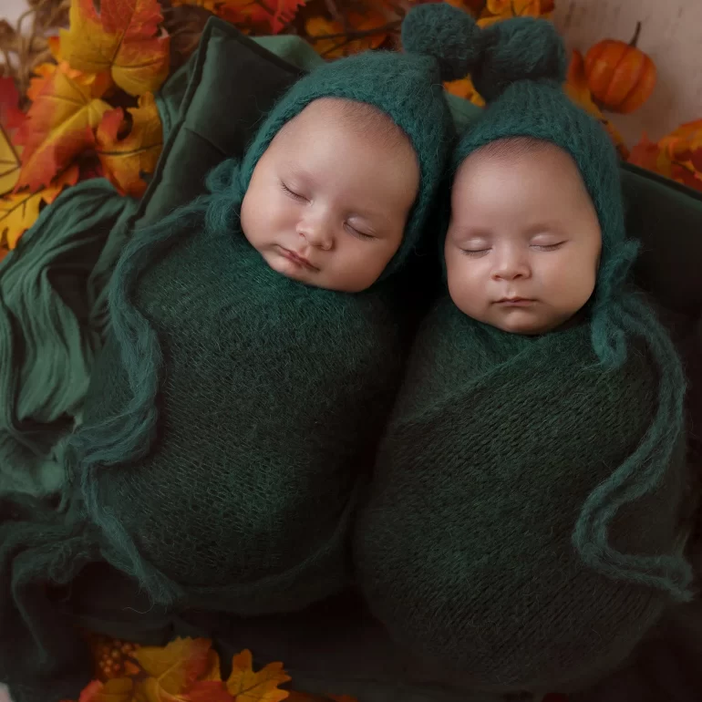 Newborn Photoshoot in North West London 85 Twin babies in forest green with pom‑pom bonnets among autumn leaves.