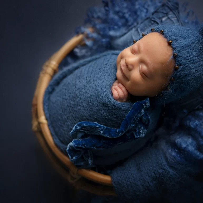 Newborn Photoshoot in North West London 12 Baby in deep blue knit and pom‑pom bonnet, asleep in wicker basket.
