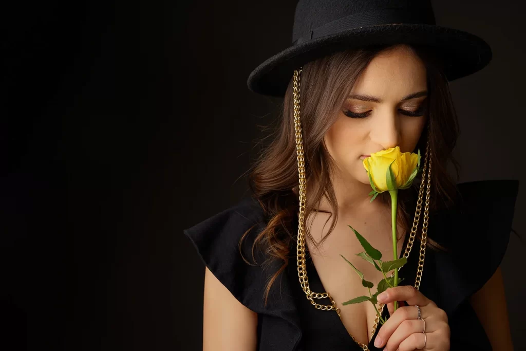 Portrait photoshoot with a woman in a black hat holding a yellow rose against a dark background.