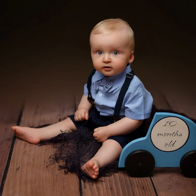 Family & Portrait Photoshoot in North West London 29 10‑month‑old boy in suspenders on rustic floor beside blue “10 months old” car prop.