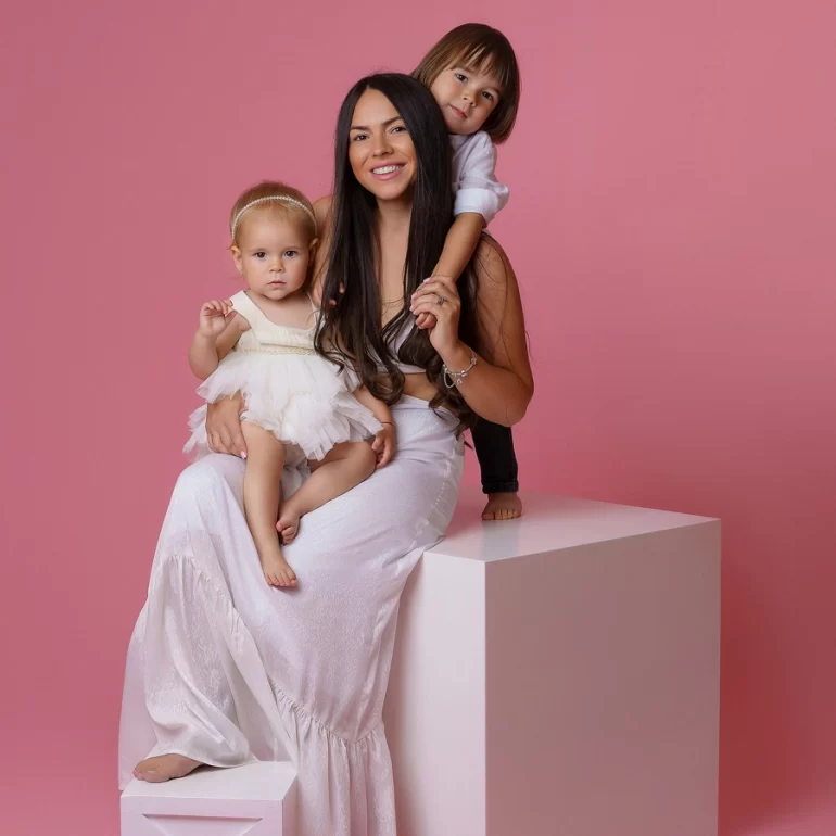Family & Portrait Photoshoot in North West London 5 Mother in white with two children; one on lap, one hugging from behind; bright pink background.