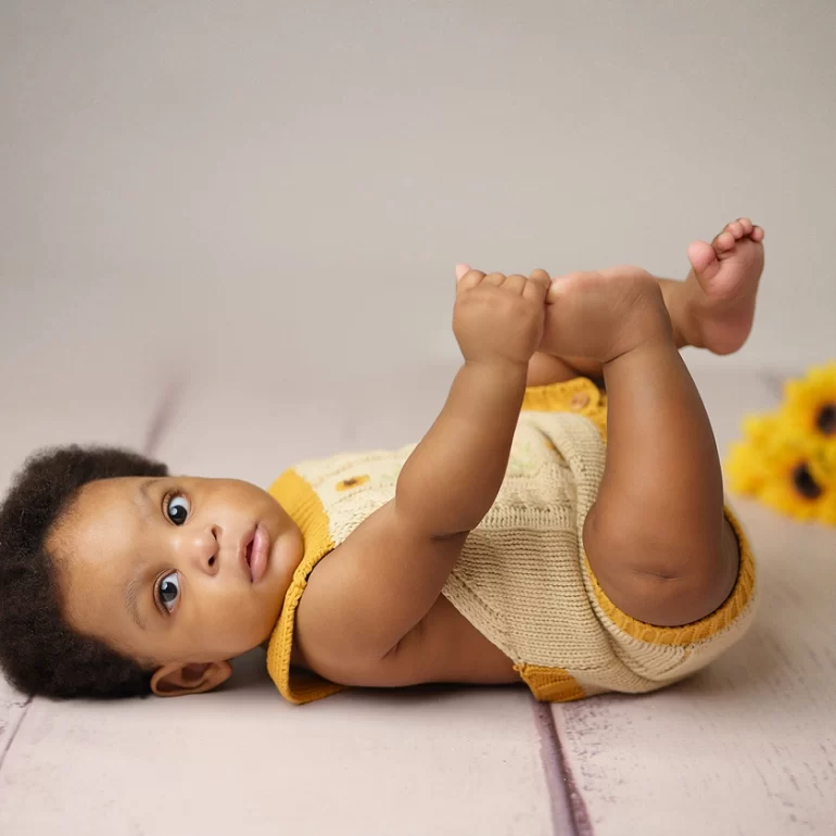 Family & Portrait Photoshoot in North West London 47 Baby in beige‑yellow knit romper on wooden floor holding feet; sunflowers behind.