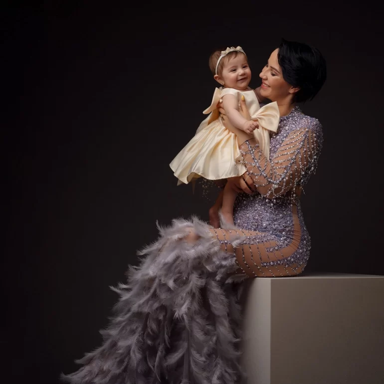 Family & Portrait Photoshoot in North West London 37 Mother in embellished gown holding smiling baby in cream dress; dark, elegant studio.