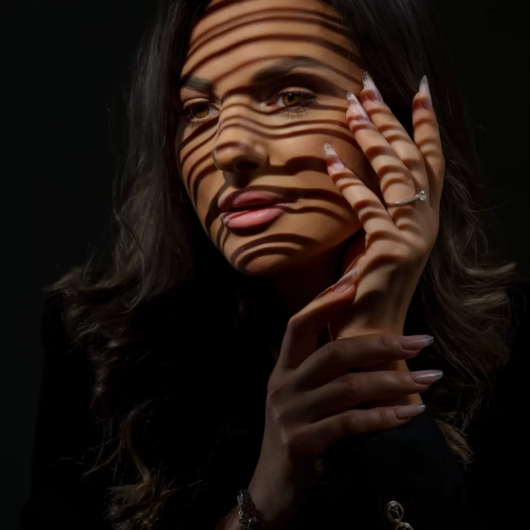Family & Portrait Photoshoot in North West London 35 Woman with soft waves under striped shadow lighting touching her face; dark moody backdrop.