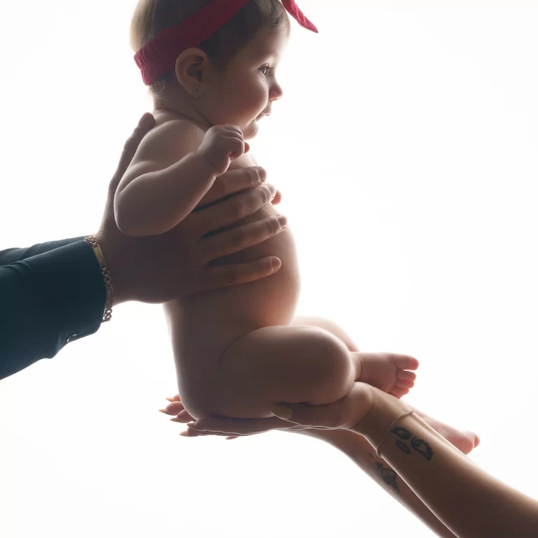 Family & Portrait Photoshoot in North West London 44 Baby in red headband lifted by adult hands against bright white background; tender silhouette.