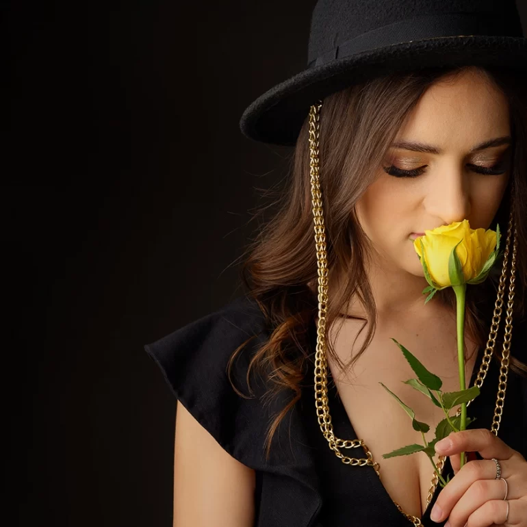 Family & Portrait Photoshoot in North West London 63 Woman in black with chain‑trim hat smelling a yellow rose; dark studio background.