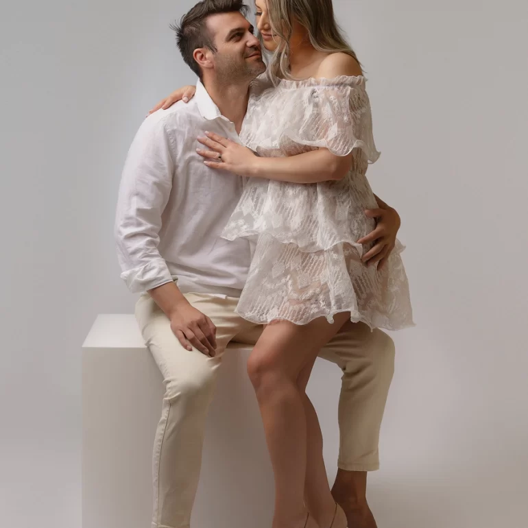 Family & Portrait Photoshoot in North West London 7 Woman in lace off‑shoulder dress seated on partner’s lap; light neutral studio scene.