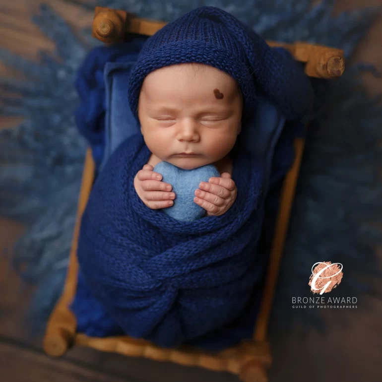 Newborn Photoshoot in North West London 1 Baby on mini wooden bed in blue knit, holding felt heart; calm studio.